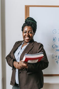 Happy black female teacher standing with workbooks near whiteboard 