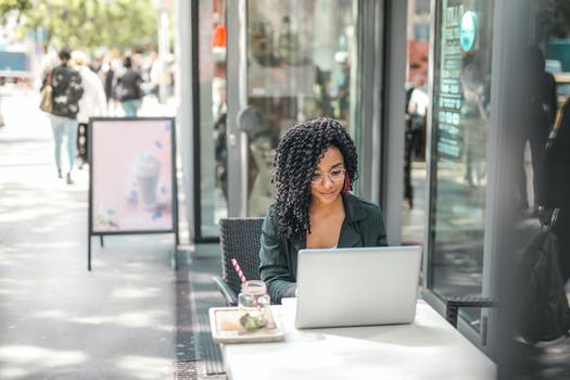 Ethnic young woman using laptop while having tasty beverage in modern street cafe